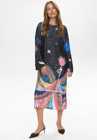 Mujer vestida con un vestido midi de manga larga con patrones abstractos y celestiales coloridos, de pie en unos tacones de esparto negros de punta afilada sobre un fondo blanco.