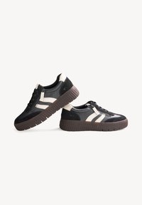 Paire de baskets basses noir et blanc avec semelles épaisses marron, fermetures éclair latérales et rayures blanches, présentées sur un fond uni.