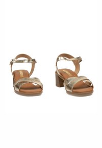 Sandalias de tacón metálico dorado con tiras entrecruzadas, correa de tobillo ajustable y una plantilla acolchada marrón. Diseño de tacón cuadrado.