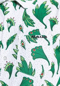 Camisa de manga corta con base blanca, que presenta un patrón de hojas verdes y acentos rojos. Incluye un bolsillo en el pecho y cierres de botón.