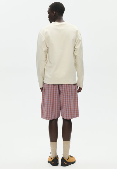 Homme vu de dos portant une chemise beige à manches longues, un short à carreaux rouge et bleu, des chaussettes beiges et des chaussures de sport jaune et noir.