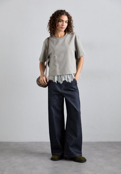 Un haut gris à manches courtes superposé sur un bas en dentelle gris clair, associé à un pantalon denim large et foncé et des chaussures vert olive.