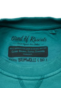 Lähikuva teal-värisen paidan kauluksesta, jossa on painettu merkki „Band of Rascals, 100 % puuvillaa (luomu)” sekä luomusertifiointitiedot.