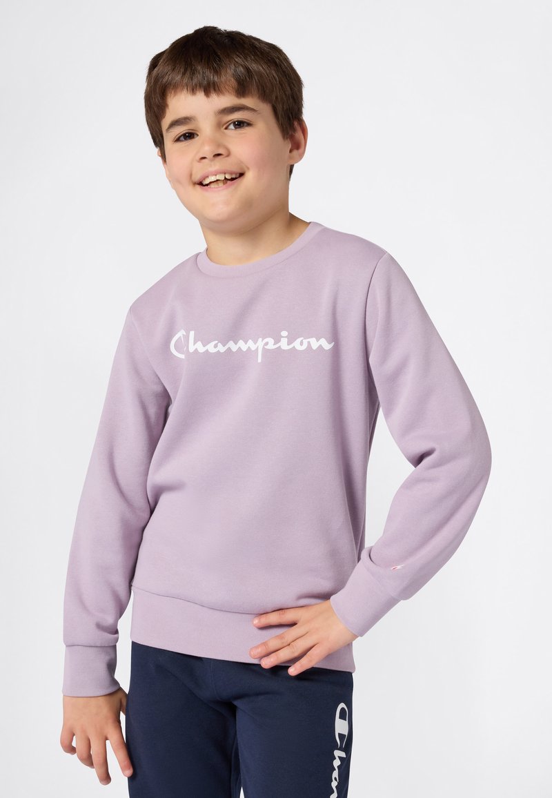 Niño sonriente que lleva un sudadera de Champion color lavanda y pantalones marinos con el logotipo de Champion, posando con una mano en la cadera frente a un fondo liso.
