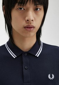 Polo shirt blu scuro con colletto a righe bianche, pattina con due bottoni e logo ricamato sul lato sinistro del petto. Texture liscia.