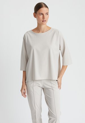 Frau mit braunen Haaren im Dutt, die ein lockeres hellbeiges Oberteil mit dreiviertel Ärmeln und passende, enganliegende Hosen vor einem einfarbigen weißen Hintergrund trägt.
