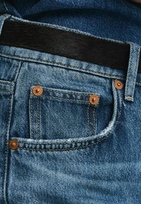 Blauwe spijkerbroek met een achterzak met koperen klinknagels. Gewatteerde stof met een lichte vervaging en een donkere riem boven de tailleband.