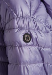 Lila Puffjacke mit gestepptem Design, glänzender Oberfläche und einem runden schwarzen Logo-Patch auf der linken Seite.
