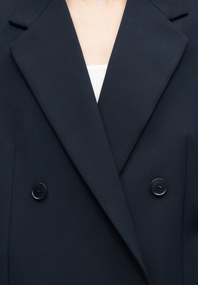 Μπλε μπλέιζερ διπλής σειράς με φαρδιές πέτες, φορεμένο πάνω από λευκό ριμπ μπλουζάκι, εμφανιζόμενο από τον λαιμό μέχρι το μέσο του κορμού.