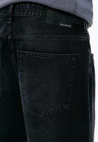 Nærbillede af en sort denimjeans baglomme med synlige syninger og Pull&Bear mærkeetiket over lommen.