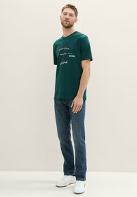 Grøn bomulds t-shirt med korte ærmer, der har trykt tekst. Båret med blå jeans og hvide sneakers for et afslappet look.