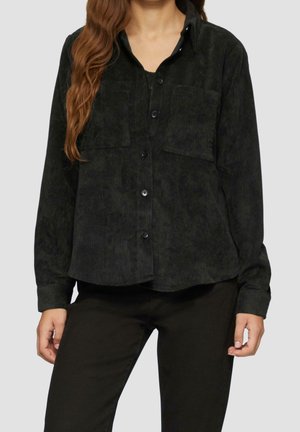 Kvinde iført en sort kordfløjls-skjorte med knapper og to brystlommer samt sorte bukser foran en ensfarvet baggrund.