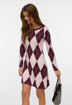 Woman wearing a long-sleeve purple and light pink argyle patterned dress, holding a black handbag, looking down and smiling.