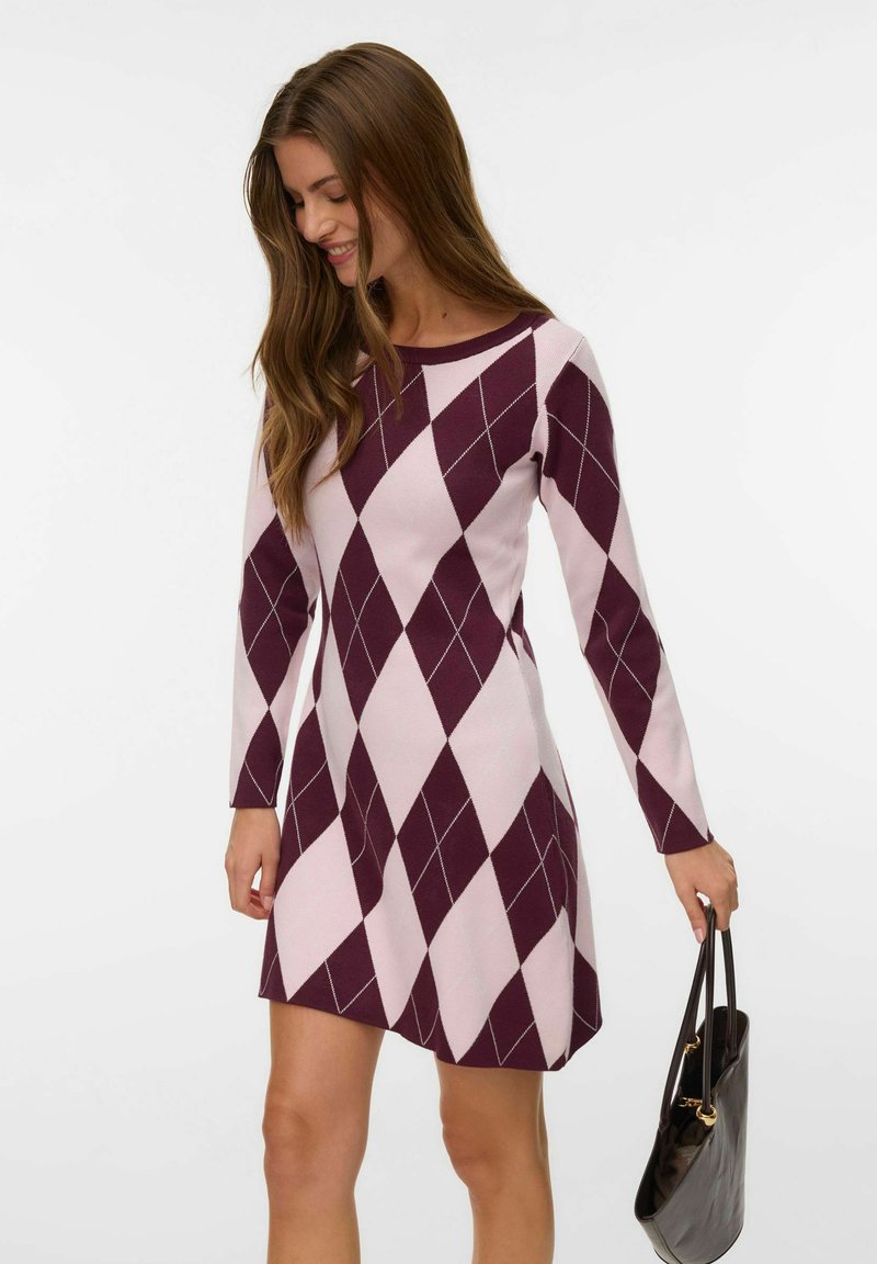 Donna che indossa un vestito a maniche lunghe con motivo argyle violetto e rosa chiaro, tiene in mano una borsa nera, guarda in basso e sorride.