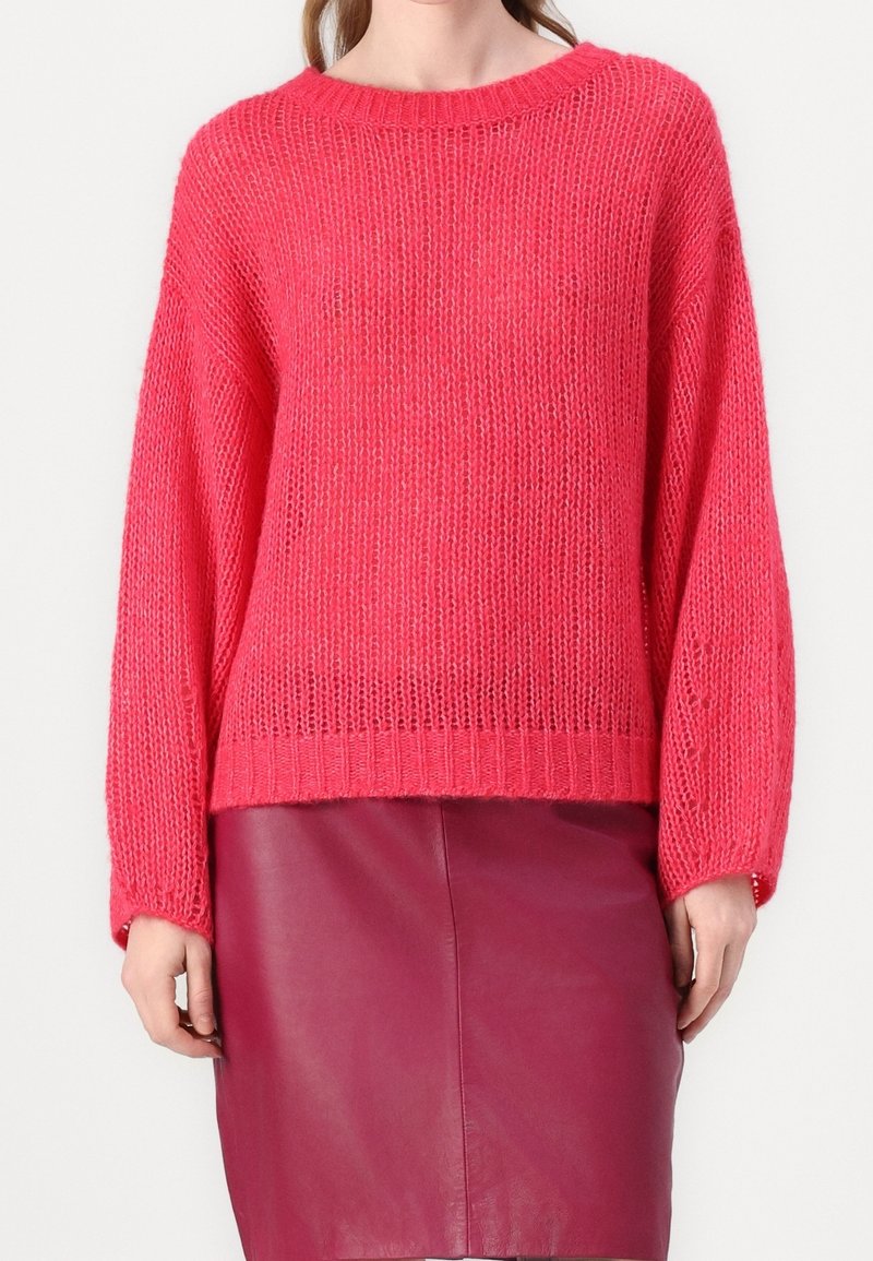 Femme portant un pull ample en tricot rose vif associé à une jupe en cuir rose foncé taille haute sur un fond uni.