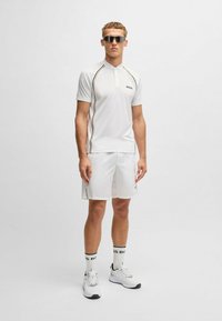 Camisa polo blanca con franjas laterales en negro y dorado, combinada con pantalones cortos blancos y zapatillas deportivas con acentos negros. Incluye gafas de sol y calcetines de marca.