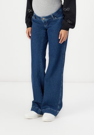 Osoba w szerokich niebieskich dżinsach, białych trampkach, szarym topie i czarnych długich rękawach z haftowanymi ozdobami w kształcie kokard.