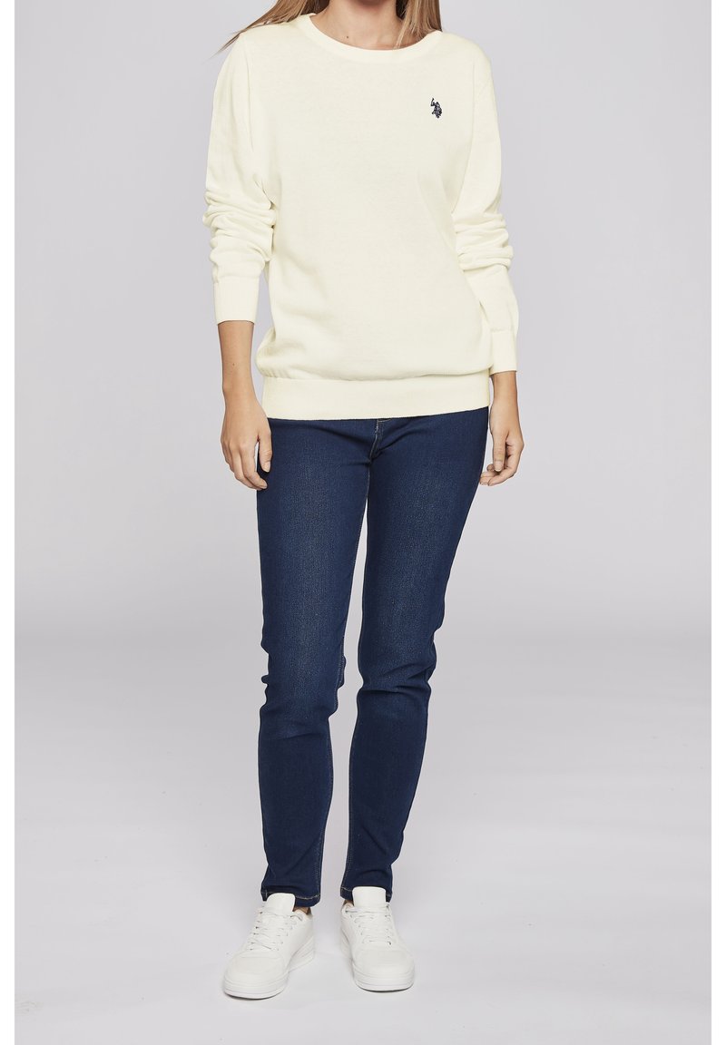 Cremefarbener Strickpullover mit Rundhalsausschnitt, gerippten Bündchen und Saum, kombiniert mit dunkelblauen Slim-Fit-Jeans und weißen Sneakers. Kleines Logo auf dem Pullover.