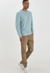 Lichtblauwe crewneck sweatshirt, tan tapered pants en witte sneakers. De sweatshirt heeft ribgebreide boorden en onderkant; de broek heeft elastische boorden.