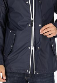Marineblaue wasserdichte Jacke mit silbernem Reißverschluss und Beschlägen, ausgestattet mit zwei Seitentaschen und einem glatten, strukturierten Stoff.