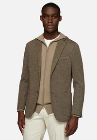 Brown textured blazer over a beige zip-up hoodie. The blazer has a single chest pocket and two front pockets. Light-colored pants.