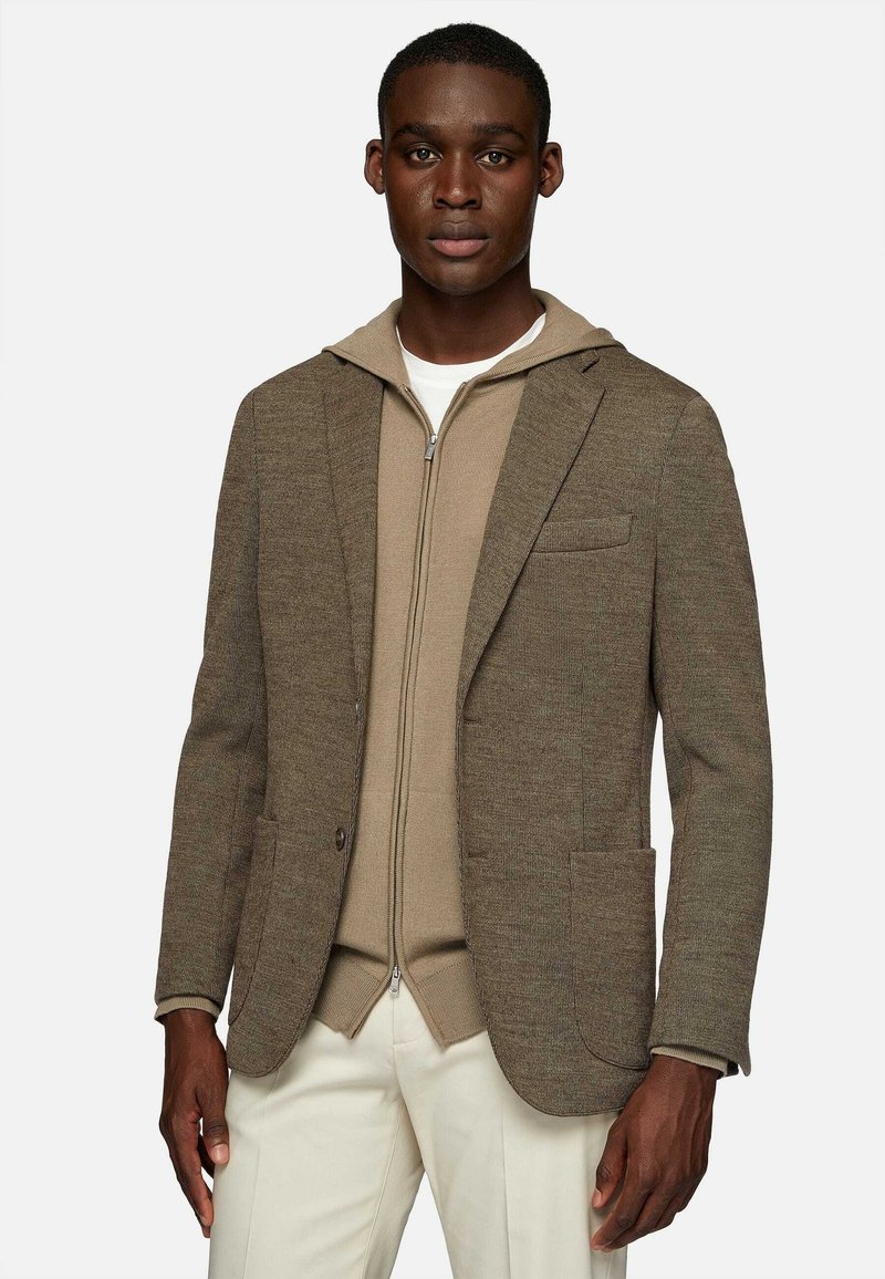Brown textured blazer over a beige zip-up hoodie. The blazer has a single chest pocket and two front pockets. Light-colored pants.