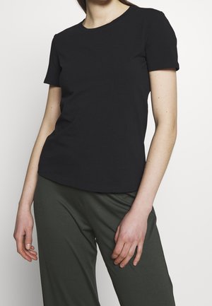 Osoba ubrana w prostą czarną koszulkę z krótkim rękawem i ciemnoszare spodnie, stojąca na białym tle z rozluźnionymi ramionami.