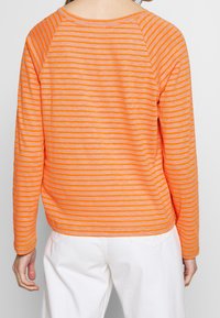 Långärmad topp i orange med växelvis tunna ränder. Har rund halsringning och raglanärmar. Materialet verkar lätt och texturerat.