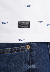 Camiseta blanca de algodón con un patrón de gafas de sol azules y una etiqueta de tela negra con texto blanco. Combinada con unos jeans de mezclilla azules.