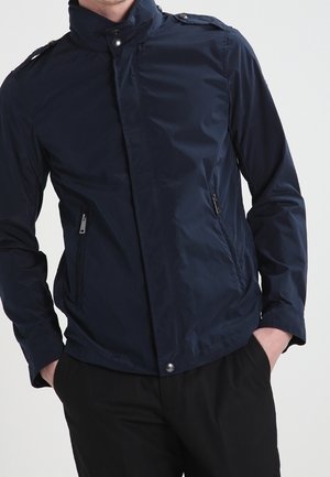 Mies pukeutuneena tummansiniseen vetoketjulliseen takkiin, jossa on nepparikiinnitteinen kaulus ja vetoketjulliset sivutaskut, kädet mustien housujen taskuissa.