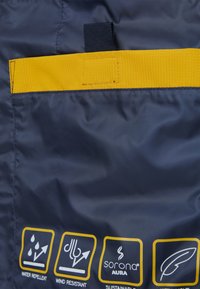 Wasserabweisender marineblauer Stoff mit gelben Akzenten. Verfügt über einen Taschenriegel und Symbole, die Windresistenz und Nachhaltigkeit anzeigen.