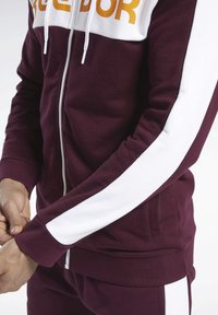 Giacca zip-up bordeaux con dettagli bianchi sulle maniche, caratterizzata da un logo grafico giallo. Tessuto morbido con polsini e vita a costine.