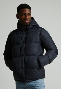 Chaqueta de abrigo azul marino con un patrón geométrico texturizado, que incluye una capucha, bolsillos laterales con cremallera y un cierre frontal abotonado.