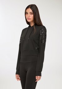Giovane donna con capelli lunghi che indossa un felpa nera con maniche decorate e leggings neri, in piedi su uno sfondo bianco semplice.