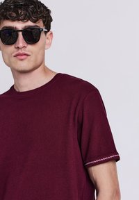 Giovane uomo con occhiali da sole scuri e una maglietta a maniche corte color bordeaux con cuciture a zigzag bianche sul bordo delle maniche, su uno sfondo chiaro.