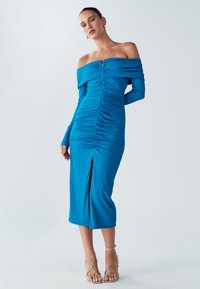 Robe bleue décolletée avec des détails froncés, manches longues et fente à l'avant. Tissu texturé avec une silhouette ajustée.