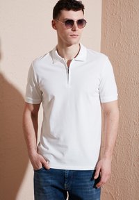 Vit pikétröja med dragkedja i kragen, korta ärmar och texturerade detaljer vid axlarna; kombinerad med mörkblå jeans.