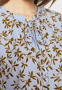 Blusa azzurro chiaro con motivo floreale verde scuro, colletto rotondo, dettagli con lacci e tessuto morbido.