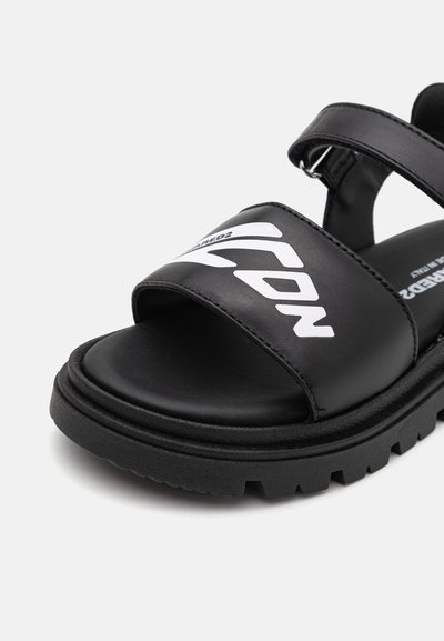 Melnas sandales ar plašu, gludu siksnu, kurai ir balts logo. Masīva melna zole ar teksturētu saķeri un nelielu izliecienu atbalstam. Minimalistisks dizains.
