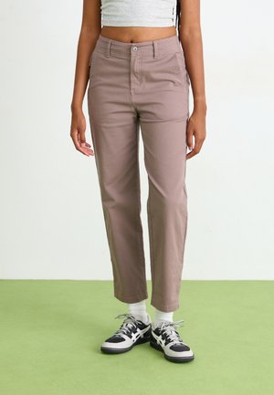 Chinos marron clair en coton avec une coupe droite. Ils présentent des poches avant et arrière, associés à des baskets noires et blanches et des chaussettes blanches.