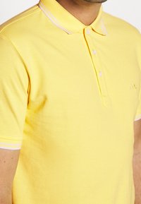 Keltainen puuvillapaita, jossa on kolmen napin napitus, ribattu kaulus vaaleanpunaisilla yksityiskohdilla ja pieni logotunnus rinnassa.