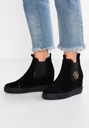 Fødder iført sorte ruskind wedge ankelstøvler med metalnitter og gyldent logo, kombineret med korte blå jeans med frynset kant.
