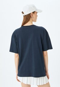 Camiseta de algodón azul marino con mangas cortas y cuello redondo, vista desde la parte trasera. Se lleva con una gorra de color claro y una falda blanca plisada.