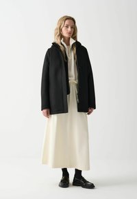 Juodas gobtuvų švarkas su lygia tekstūra, turintis užtrauktuką ir sagų uždarymą, dėvimas ant baltos ilgomis rankovėmis suknelės ir kreminės spalvos sijono.