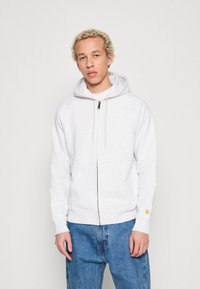 Hellgrau Zip-Hoodie aus weichem Material, mit einem vorderen Reißverschluss, zwei Seitentaschen und einem kleinen Logo auf dem linken Ärmel.