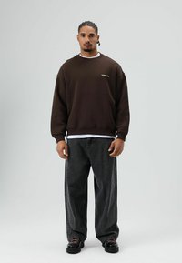 Bruine oversized sweatshirt met gouden tekst, gecombineerd met zwarte jeans met wijd uitlopende pijpen en zwarte schoenen. De stof voelt zacht aan, het ontwerp heeft een relaxte pasvorm.