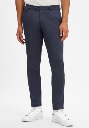 Man draagt donkerblauwe slim-fit broek en witte sneakers, staand tegen een witte achtergrond.