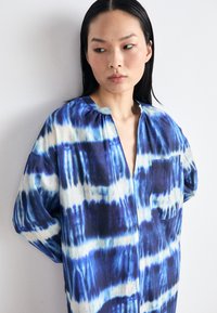 Cămașă bleu și albă cu model tie-dye, confecționată dintr-un material cu o croială lejeră, cu decolteu în V, buzunar pe piept și mâneci ușor bufante.