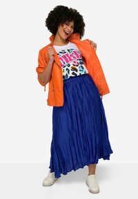 Chaqueta naranja brillante, camiseta gráfica blanca con estampado de animales colorido y una falda azul fluida. Texturas suaves con zapatillas blancas casuales.
