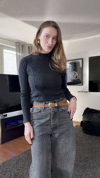 Top preto canelado de mangas compridas combinado com jeans cinzentos de cintura alta. Cinto castanho com fivela prateada; cabelo longo e ondulado e unhas escuras.